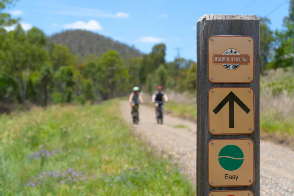 Two cyclists ride on a dirt pathway surrounded by greenery, with a signpost indicating the 