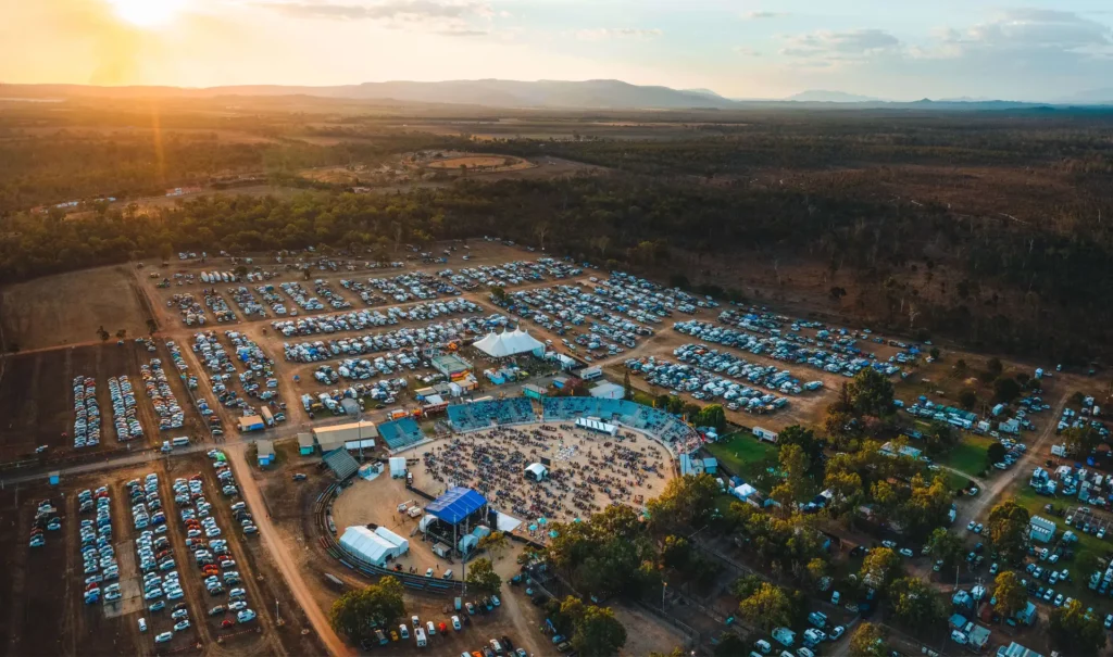 Aerial view of an outdoor event at sunset, with rows of parked cars surrounding a central area with tents, a stage, and people gathered, set in a vast, rural landscape.