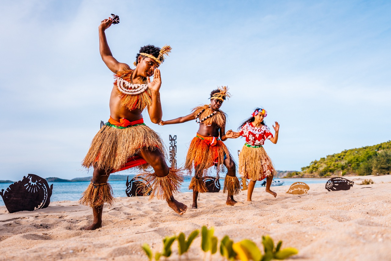 Three people are performing a traditional dance on a sandy beach, dressed in grass skirts and adorned with headpieces, against a backdrop of the sea and distant greenery.