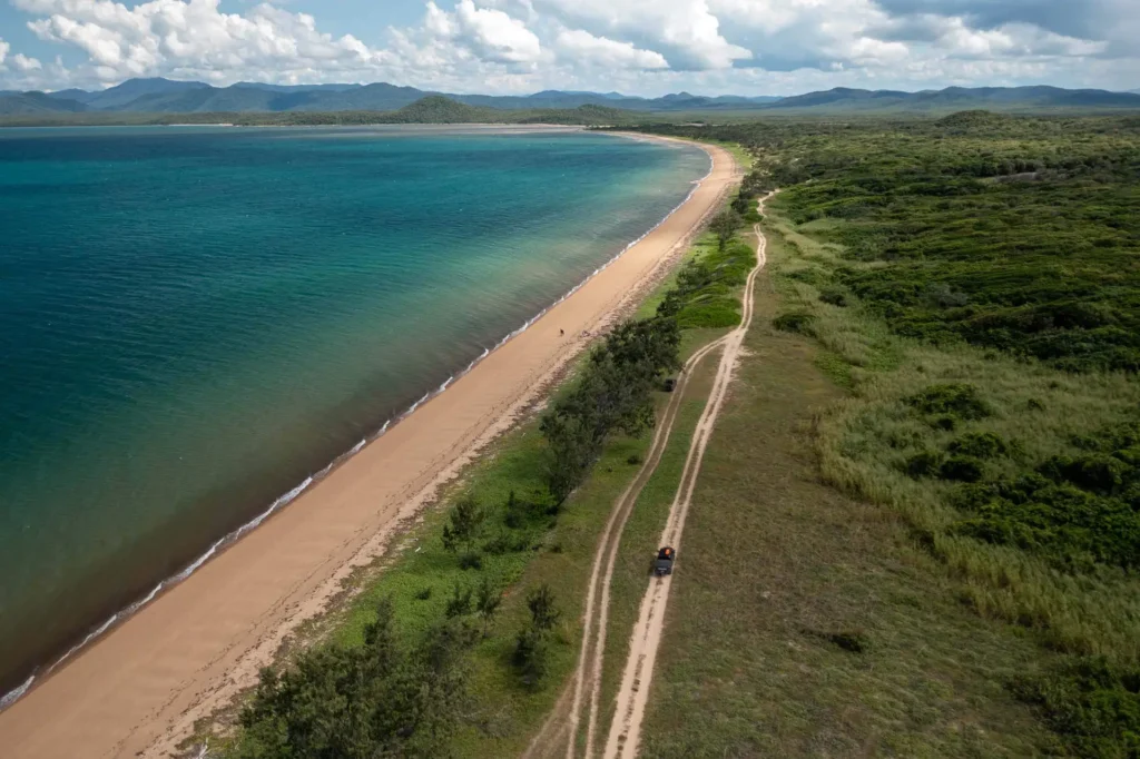 Aerial view of a long sandy beach with clear blue water on the left and a dirt road running parallel to the shoreline on the right, surrounded by lush greenery and mountains in the distance.