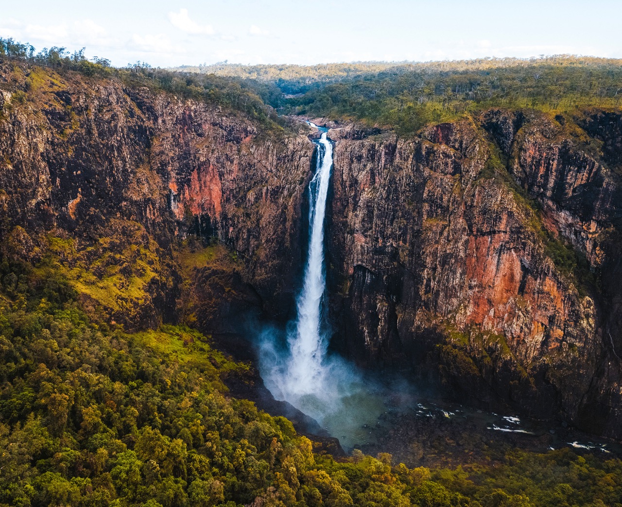 A tall waterfall cascades down rugged, rocky cliffs surrounded by dense forest, with mist rising from the base and a river flowing away.