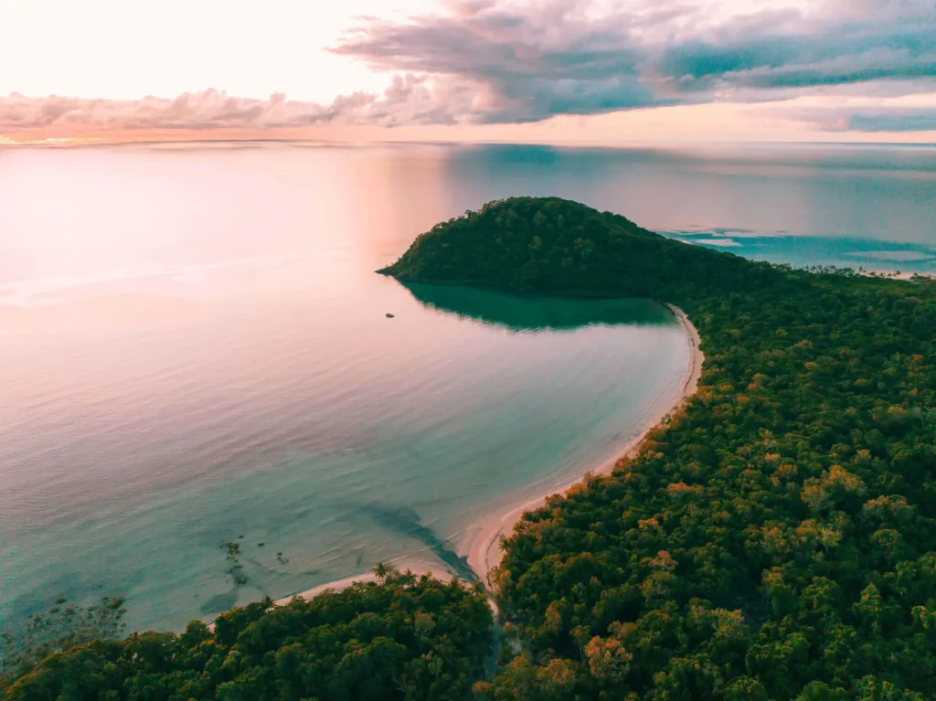 Aerial view of a coastal landscape with a lush, tree-covered hill and a curved sandy beach along calm, clear water at sunset. Cloudy skies are above the horizon.