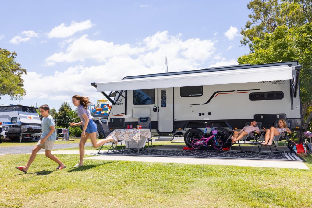 Family sitting outside a parked RV, with two children running on the grass. Bicycles and a table with a checkered cloth are nearby. Trees and a clear sky in the background.