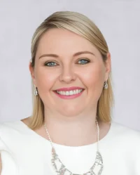 A woman with straight blond hair, wearing a white top, silver earrings, and a silver necklace, smiling at the camera against a plain light background.