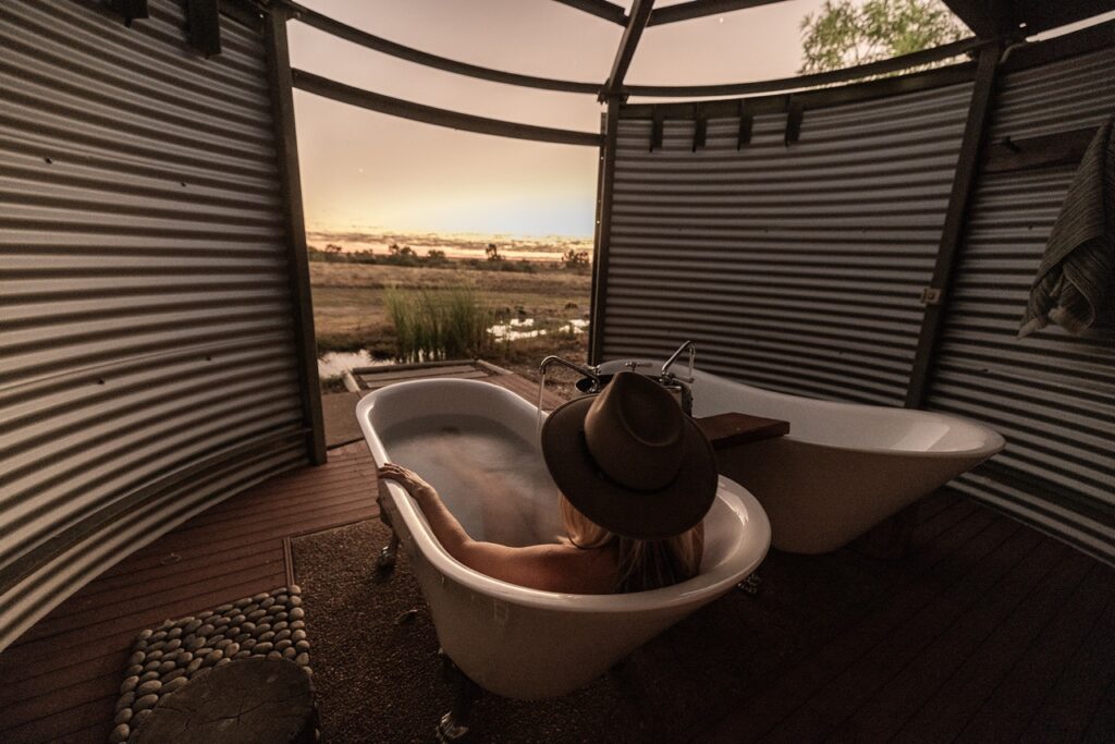 A person in a hat relaxes in a clawfoot bathtub, enjoying the view of a serene landscape through a corrugated metal enclosure. Another empty clawfoot tub is positioned nearby.