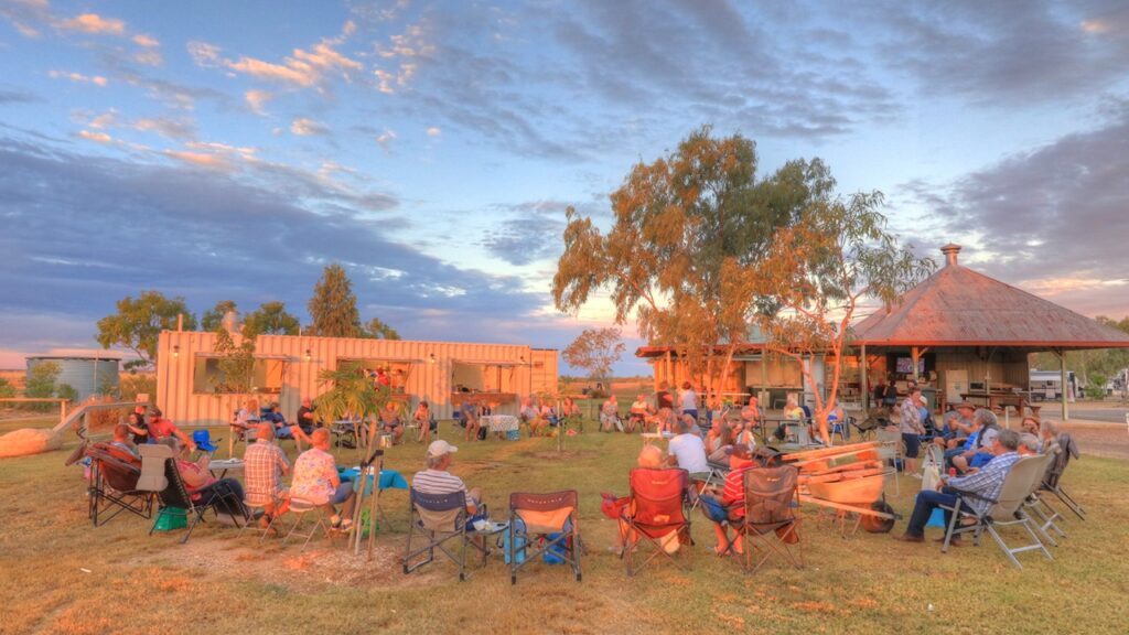 A group of people sitting in camping chairs forms a large circle outdoors, with trees and a building in the background, under a partly cloudy sky at sunset.