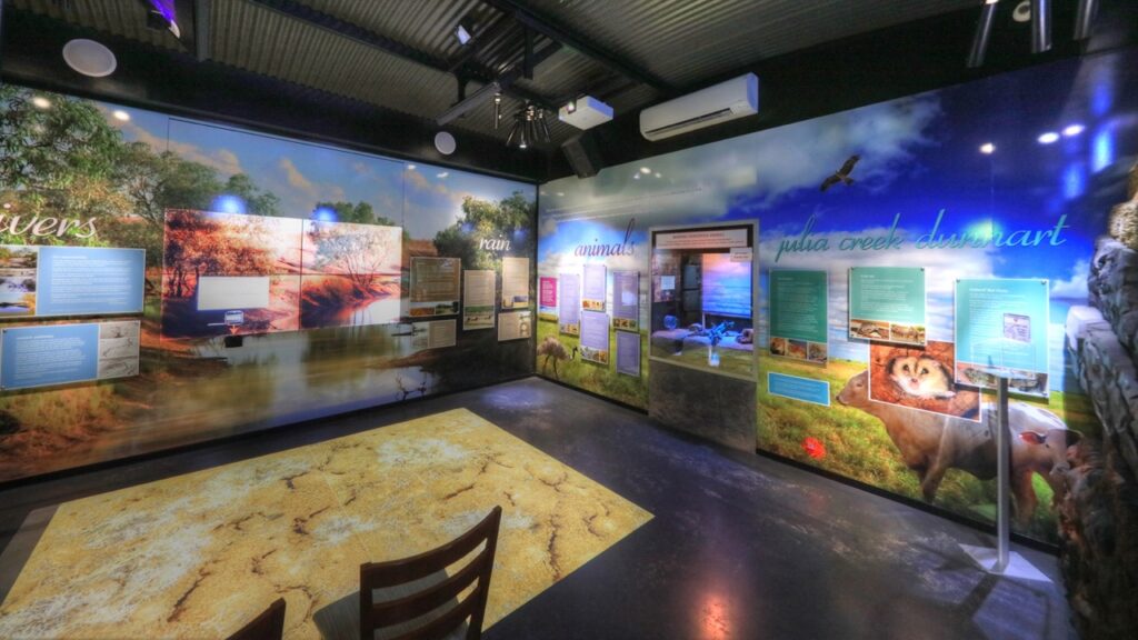 A well-lit museum exhibit displaying informational panels, photographs, and illustrations about wildlife and natural habitats. Chairs are placed in the center of the room.