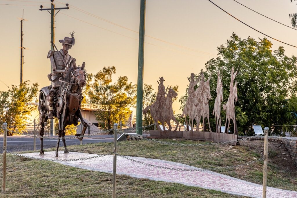 A metal sculpture of a cowboy on horseback and a herd of cattle stands in an outdoor setting, surrounded by grass and utility poles.