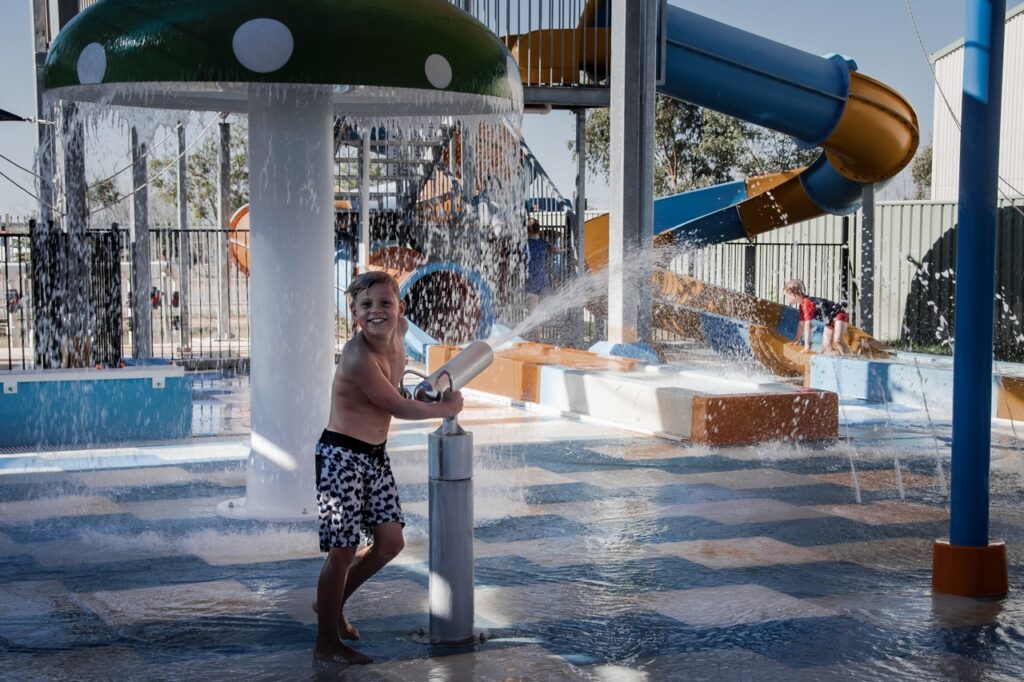 A child wearing black and white swim trunks plays with a water cannon at a water park, standing under a large mushroom-shaped water feature. Other children are seen on slides and in the background.