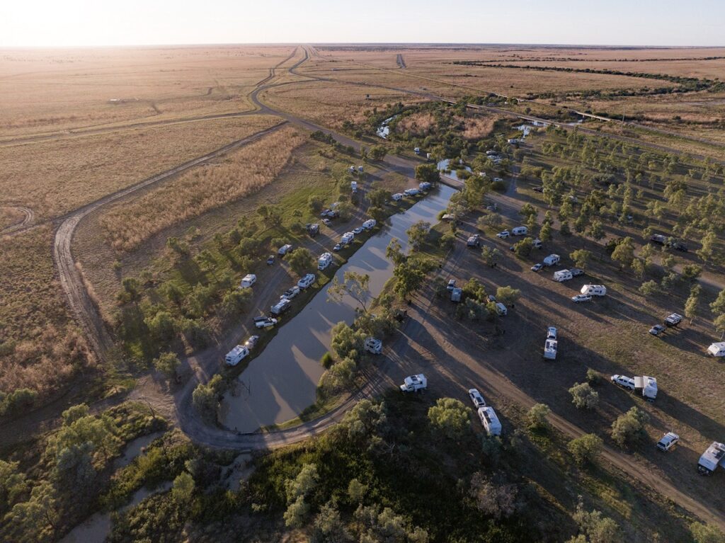 Aerial view of a campsite with numerous caravans positioned around a water body in a vast, open landscape with sparse vegetation.