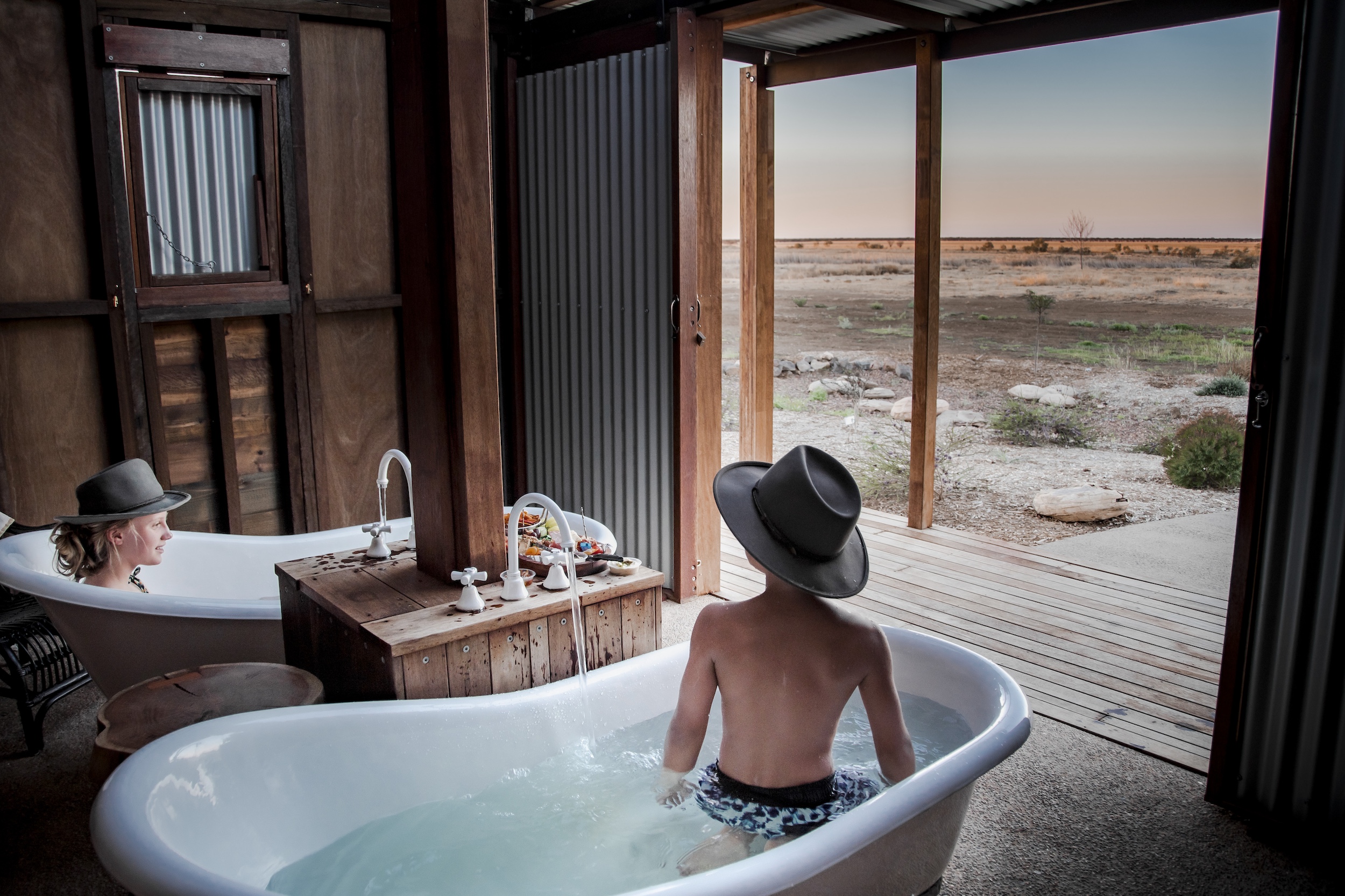 Two people sit in separate outdoor bathtubs wearing hats, looking out toward a dry landscape through an open door. The scene is set under a rustic, corrugated metal shelter.