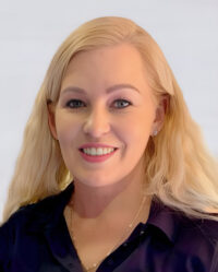 A blonde woman with long hair and blue eyes smiles at the camera. She is wearing a black shirt and a gold necklace. The background is plain and light-colored.