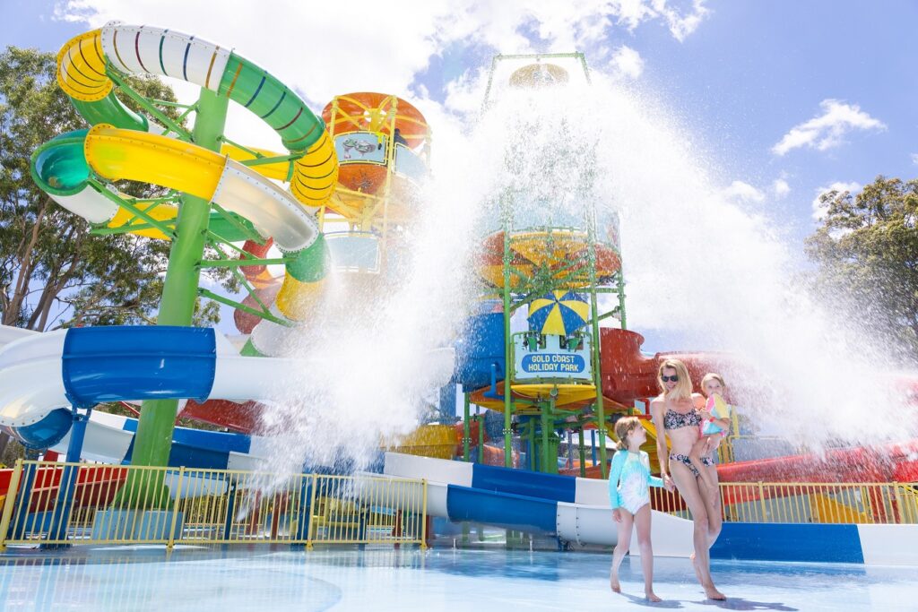 A woman and two children stand near a water slide at a water park, with large water splashes and colorful slides in the background.