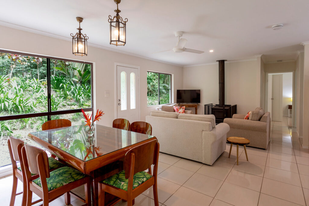 Bright living and dining area with a glass table, wooden chairs, two sofas, a TV, and large windows overlooking greenery. Two ceiling lights and a ceiling fan are visible.