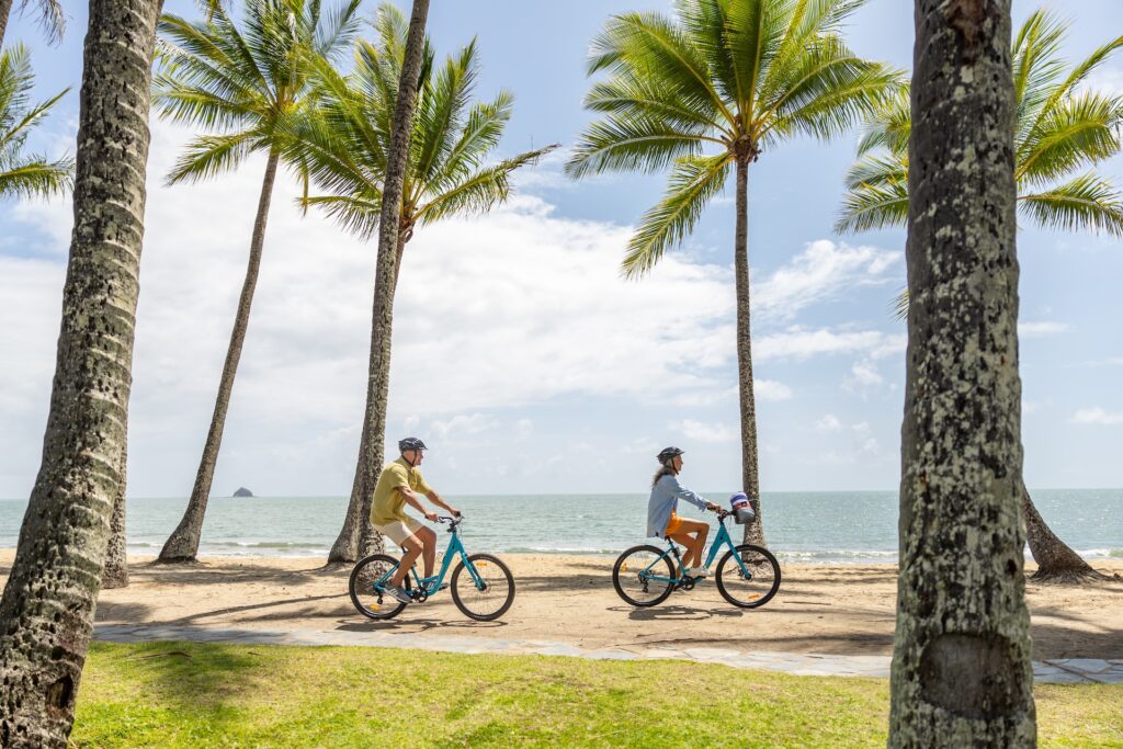 Two people ride bicycles along a sandy beach path lined with palm trees, with the ocean and a partly cloudy sky in the background.