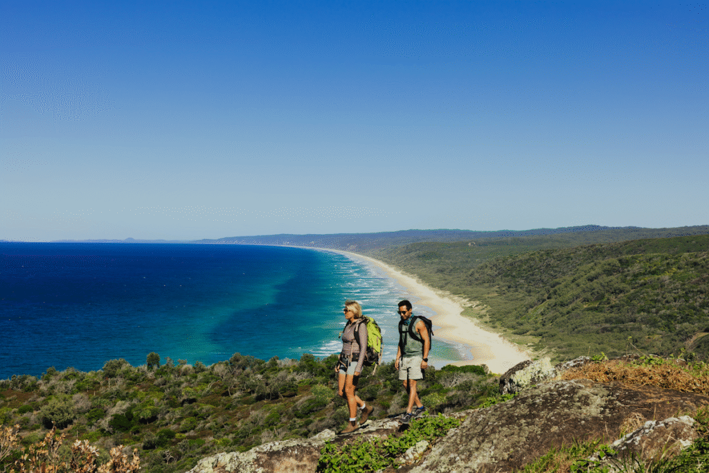 Two people with backpacks hike along a coastal trail overlooking a long sandy beach, blue ocean, and forested landscape under a clear sky.