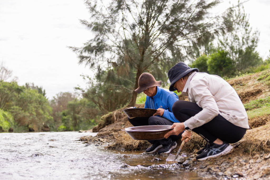 Two people in hats crouch by a riverbank, panning for materials in shallow water with large shallow pans, surrounded by trees and greenery.