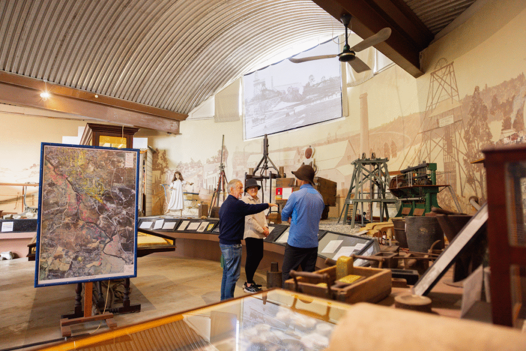 Three people stand inside a museum exhibit room with historical artifacts, display cases, maps, and a large wall mural depicting an industrial scene.