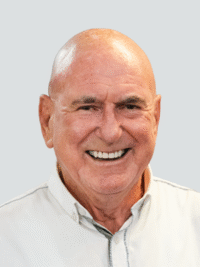 A smiling older man with a bald head wearing a white collared shirt, shown against a plain light gray background.