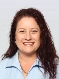 A woman with long, wavy brown hair and a blue collared shirt smiles at the camera against a plain light background.