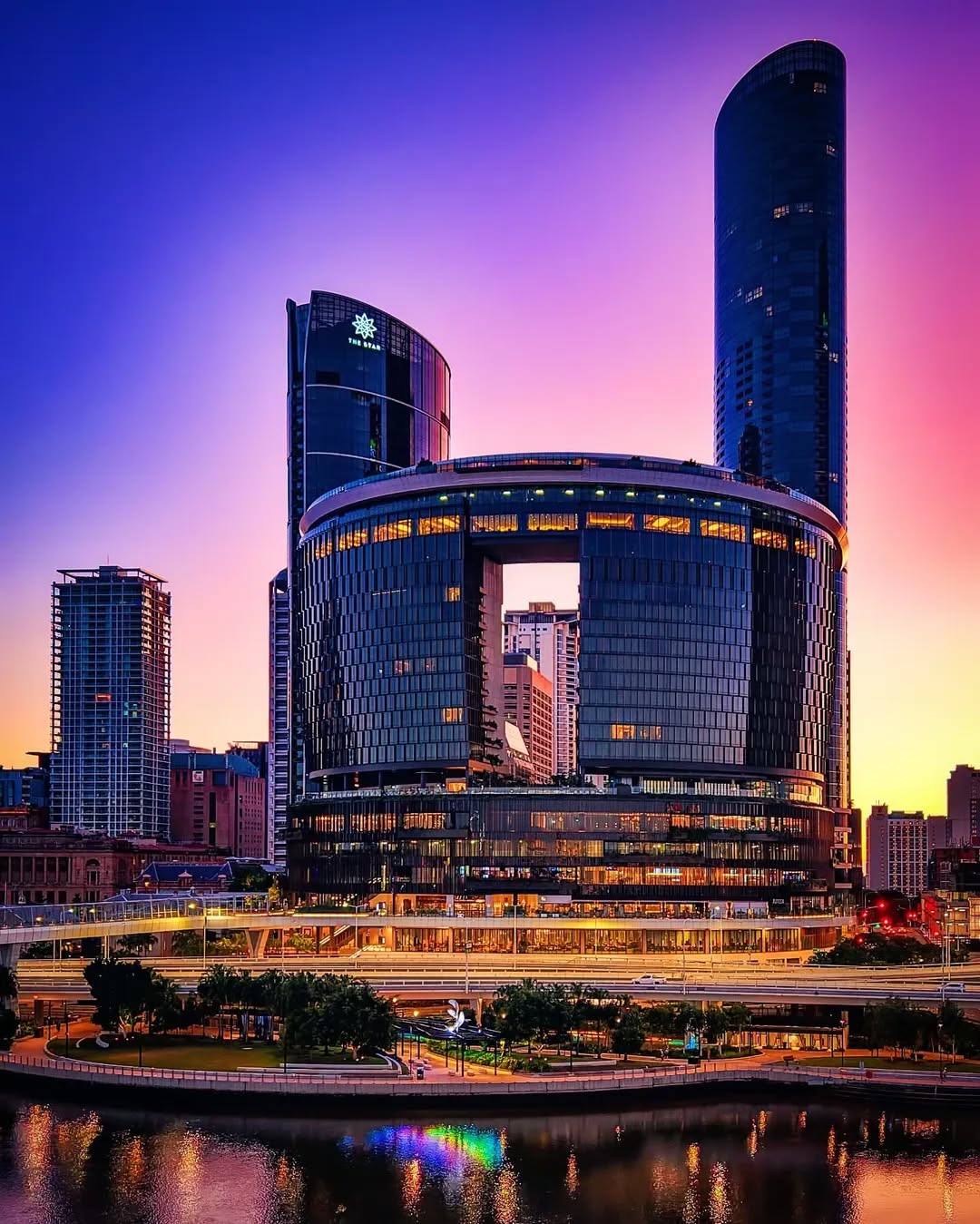 Modern high-rise buildings with glass facades reflecting colorful sunset sky, situated by a river with greenery and lights in the foreground.