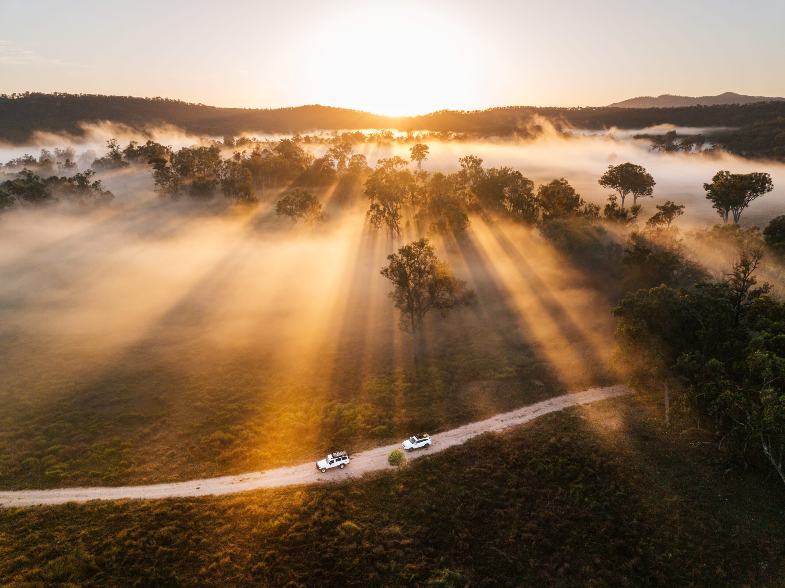 Aerial view of two white vehicles on a dirt road with sunrise, mist, and sun rays filtering through trees in a rural landscape.