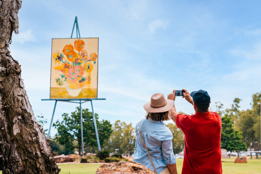 Two people stand outdoors taking a photo of a large floral painting displayed on an elevated easel, with trees and a blue sky in the background.