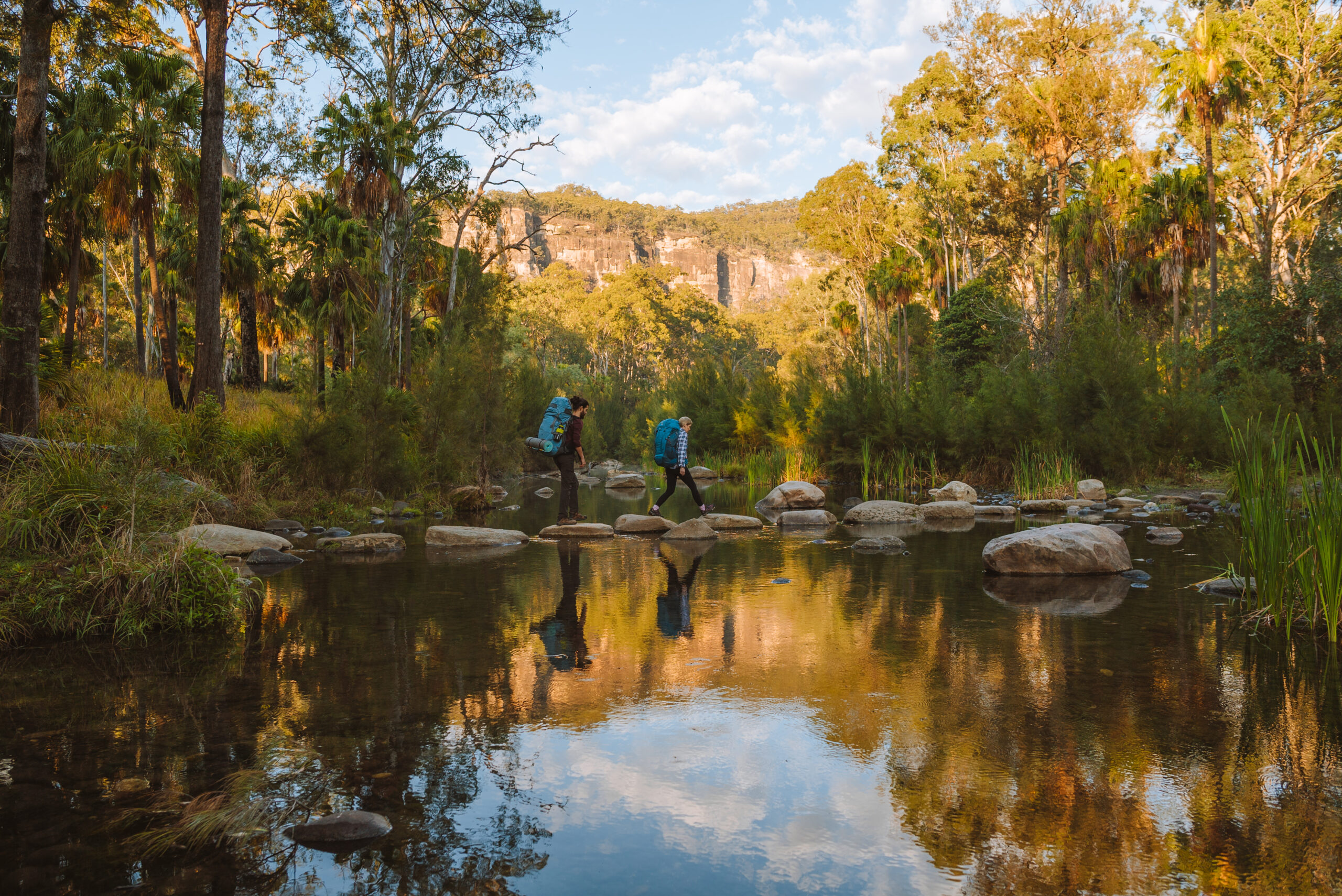 Two hikers with backpacks cross a shallow creek on stepping stones, surrounded by trees and cliffs under a partly cloudy sky.