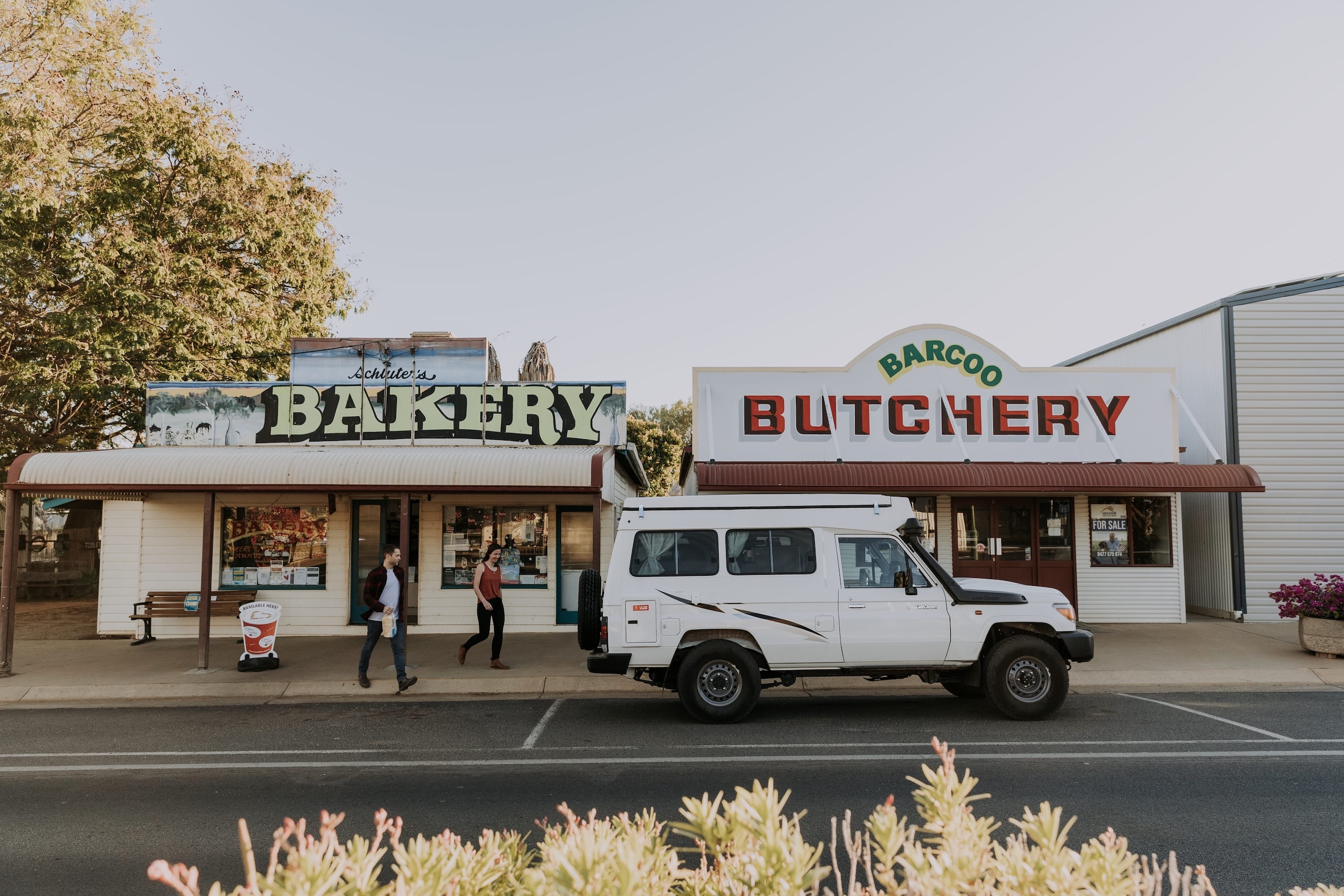 A white SUV is parked in front of a bakery and a butchery on a small town street; two people walk on the sidewalk.