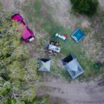 Aerial view of a campsite with a pink vehicle, two tents, and several people gathered around a picnic table on a grassy area near trees and a dirt path.
