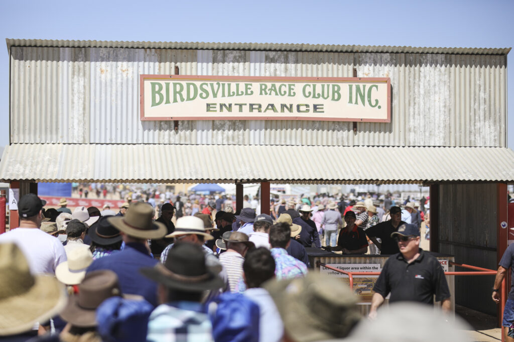 Crowd of people wearing hats enters the Birdsville Race Club under a corrugated metal sign on a sunny day.