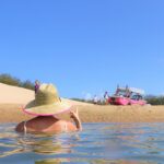 Person in a straw hat wades in shallow water near a sandy shore with a pink boat and several people in the background under a clear blue sky.