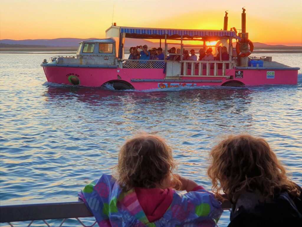 Two children watch a pink boat with passengers on the water at sunset, with distant hills visible in the background.