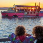 Two children watch a pink boat with passengers on the water at sunset, with distant hills visible in the background.