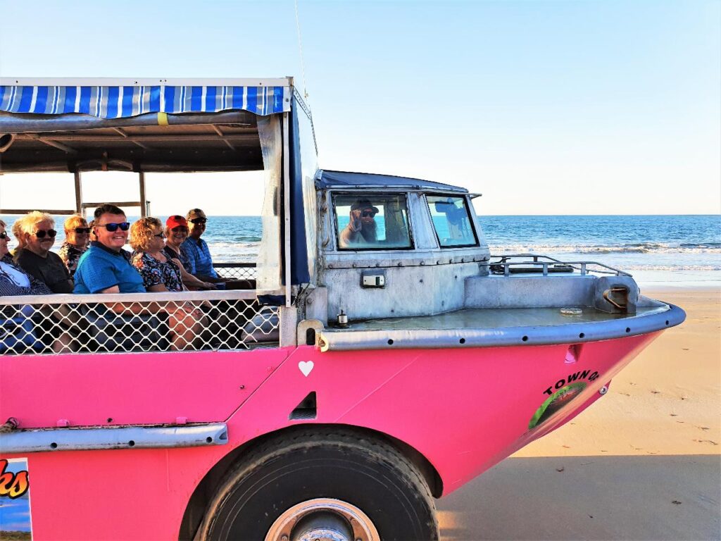 A group of people ride in a pink amphibious vehicle on a sandy beach near the ocean under a clear sky.