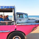 A group of people ride in a pink amphibious vehicle on a sandy beach near the ocean under a clear sky.