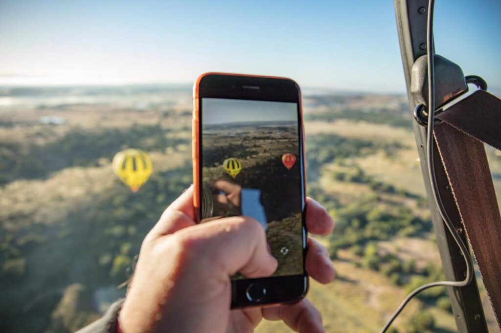 A person holds a smartphone, taking a photo of two hot air balloons floating over a landscape, as seen from another hot air balloon.