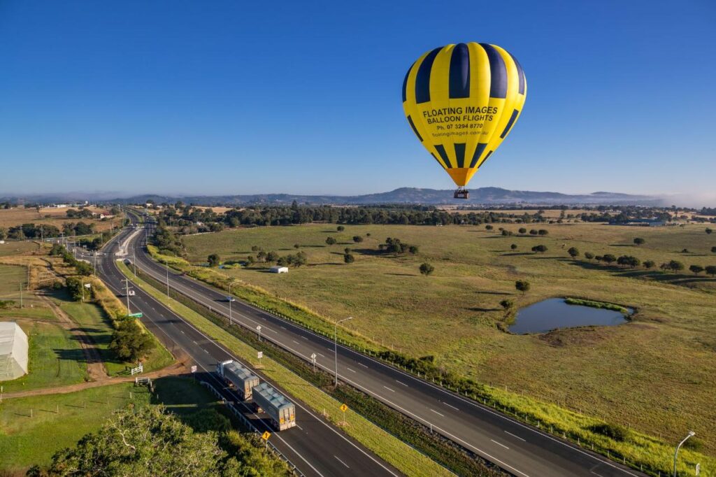 A yellow and black hot air balloon floats over a rural landscape with fields, a pond, and a highway on a clear day.