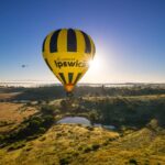 A yellow and black hot air balloon with "discover Ipswich" floats above a grassy landscape at sunrise, with a helicopter in the distance.