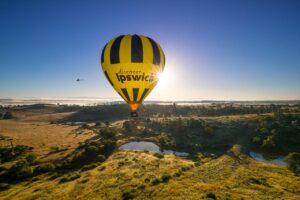A yellow and black hot air balloon with "discover Ipswich" floats above a grassy landscape at sunrise, with a helicopter in the distance.