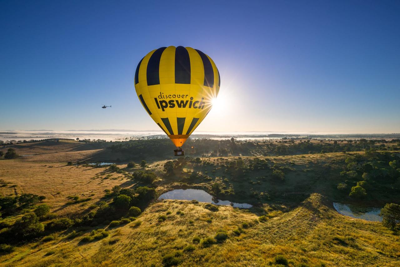 A yellow and black hot air balloon with "discover Ipswich" floats above a grassy landscape at sunrise, with a helicopter in the distance.