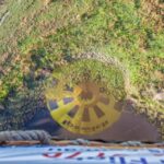 A hot air balloon casts a round yellow shadow with dark sections on a grassy and marshy area, viewed from the basket above.