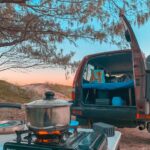 A butane stove with a pot sits on a table near a parked vehicle with its back door open, showing a bed setup inside. Trees and sky are visible in the background.