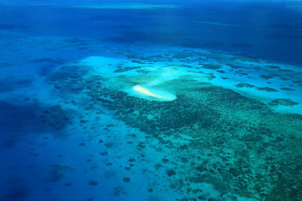 Aerial view of a small sandy island surrounded by shallow, clear blue water and coral reefs in the ocean.