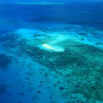 Aerial view of a small sandy island surrounded by shallow, clear blue water and coral reefs in the ocean.