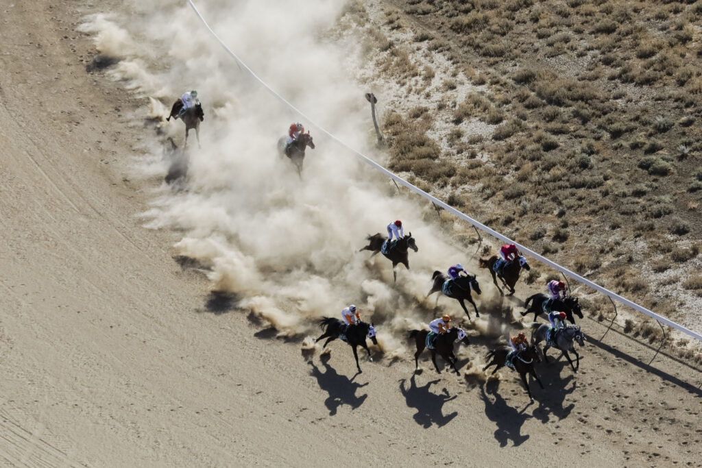 Aerial view of a group of jockeys riding horses around a dirt racetrack, kicking up clouds of dust as they race.