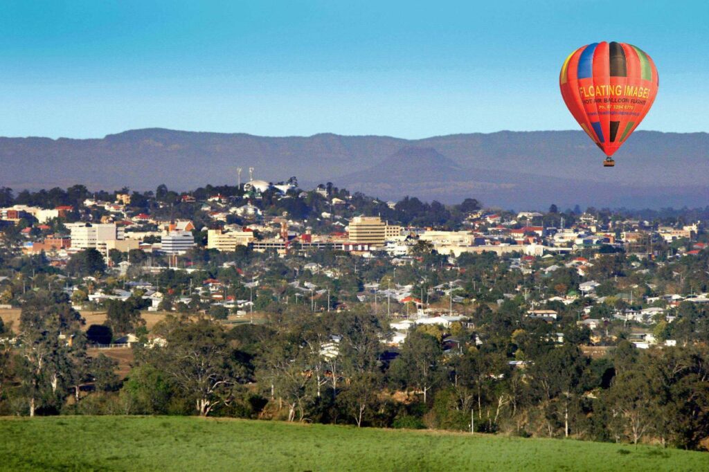 A red and orange hot air balloon floats above a town with suburban houses and buildings, set against distant hills and a clear blue sky.