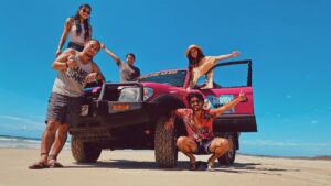 Five people pose cheerfully around a pink off-road vehicle parked on a sandy beach under a clear blue sky.