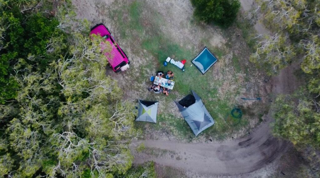 Aerial view of a campsite with three tents, a group of people sitting at a table, a pink vehicle, and surrounding trees.