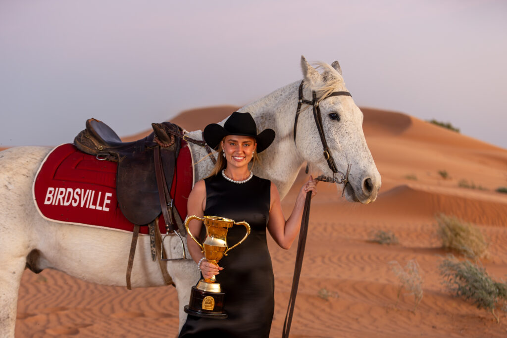 A woman in a black dress and cowboy hat holds a trophy beside a white horse with a 